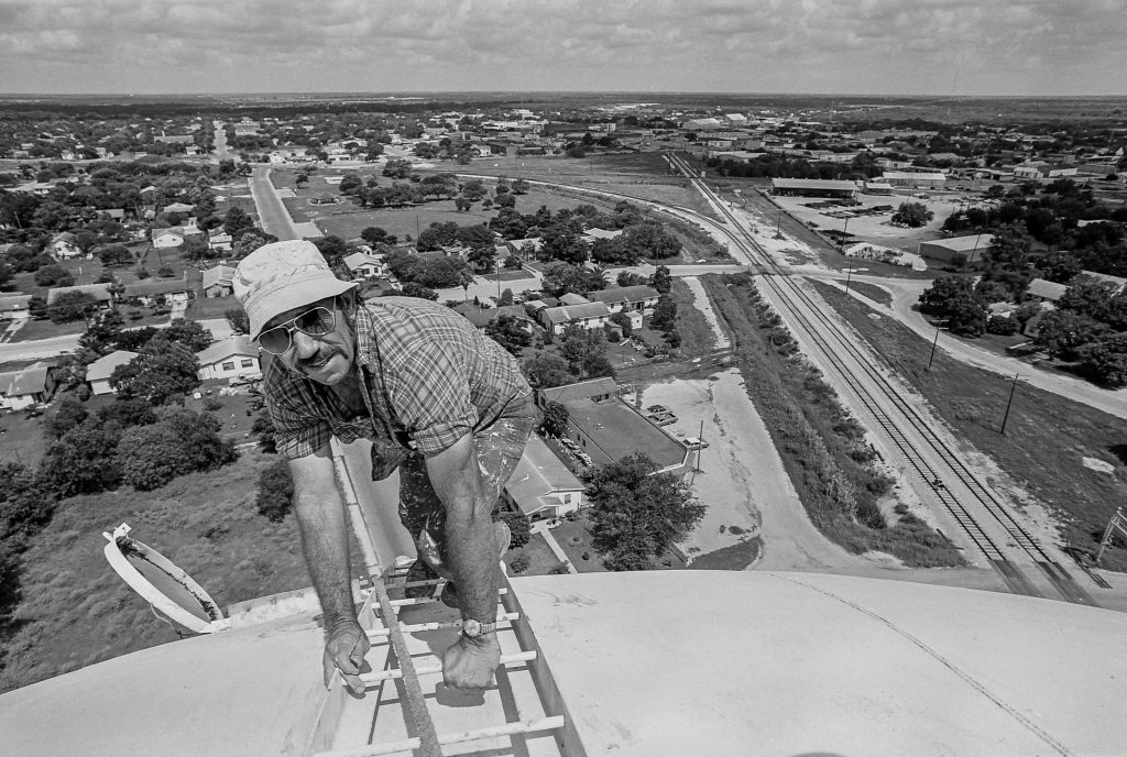 Scan10865 Photo of worker climbing tank of tall water tower with town in background below